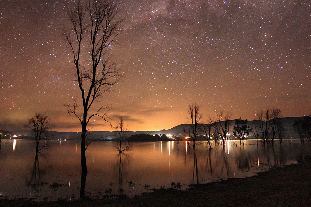 a lake at night with a starry sky and lights on the distant shore